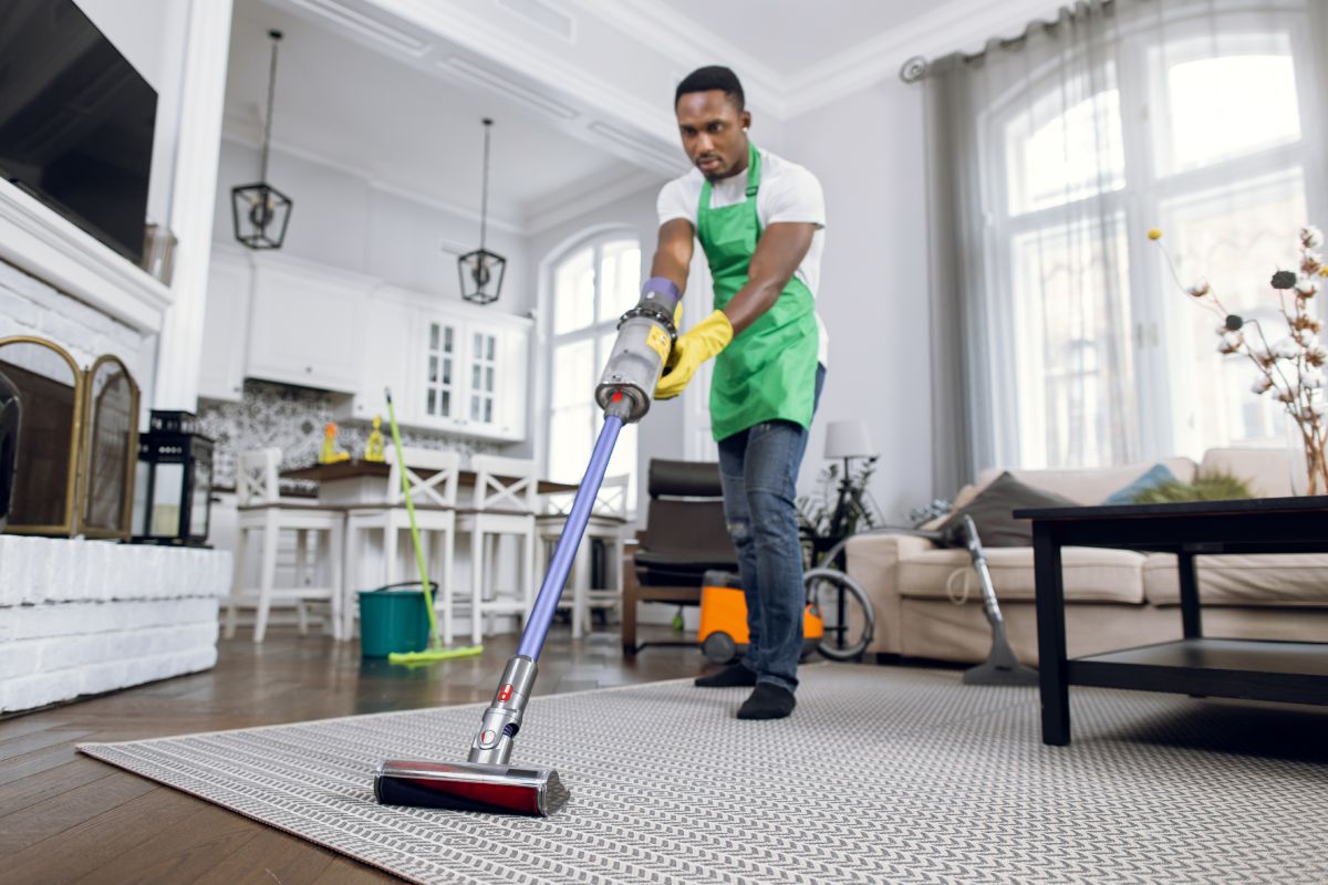African man removing dust from soft carpet with modern vacuum cleaner. Male cleaner wearing green apron and yellow rubber gloves. Working process at bright room.