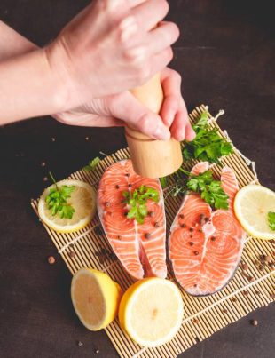 A woman sprinkles salmon with pepper from a pepper mill.