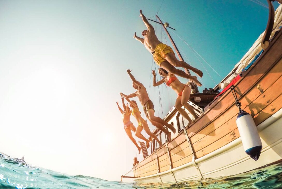 Group of happy friends diving from sailing boat into the sea - Young people jumping inside ocean in summer vacation - Main focus on right man - Travel and fun concept - Fisheye lens distortion