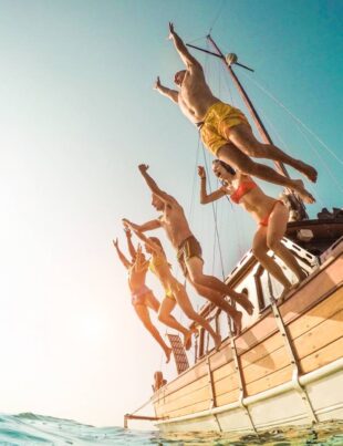 Group of happy friends diving from sailing boat into the sea - Young people jumping inside ocean in summer vacation - Main focus on right man - Travel and fun concept - Fisheye lens distortion
